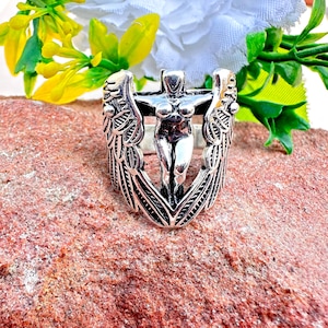 Puede incluir: Un anillo plateado con una figura femenina central en una cruz, flanqueada por alas detalladas. El anillo se encuentra sobre una superficie texturizada de color marrón rojizo, con flores amarillas y blancas en el fondo. El diseño del anillo es intrincado y detallado.