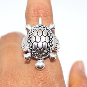 Puede incluir: Anillo de tortuga plateado. El anillo presenta un diseño detallado de tortuga con un caparazón texturizado, pequeñas patas y una cabeza. El anillo se lleva en un dedo, mostrando la intrincada artesanía y el diseño de la joya.