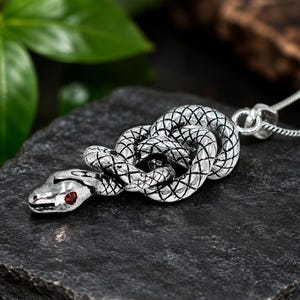 Puede incluir: Un colgante de serpiente en tono plateado con un diseño enroscado, texturas de escamas detalladas y una pequeña piedra preciosa roja incrustada en el ojo. El colgante está unido a una fina cadena de eslabones en tono plateado.