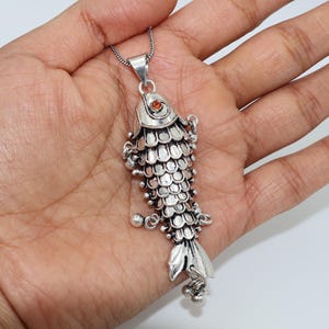 May include: A silver-colored fish pendant with a small orange gem eye, suspended from a delicate chain. The fish has detailed scale and fin designs, with small dangling accents. The pendant is held in a hand against a white background.