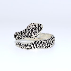 Puede incluir: Anillo de serpiente plateado con un patrón texturizado en forma de escamas. La cabeza de la serpiente está levantada y el anillo se enrolla alrededor del dedo. El anillo está sobre un fondo blanco.
