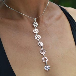 Peut inclure: Un collier pendentif argenté avec sept symboles de chakra disposés verticalement. Le collier est présenté sur un fond sombre, mettant en valeur les motifs détaillés des symboles de chakra.