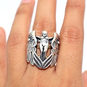 Puede incluir: Anillo plateado con una figura central con los brazos extendidos, flanqueada por diseños de alas detallados. El anillo tiene una estética vintage, con detalles intrincados en las alas y la figura. El anillo se lleva en un dedo.