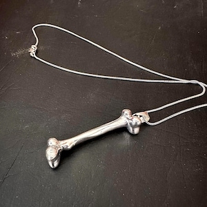 May include: A silver-colored bone pendant necklace. The pendant is a detailed replica of a bone, with a textured surface. The necklace chain is a delicate silver chain. The necklace is displayed on a dark surface.