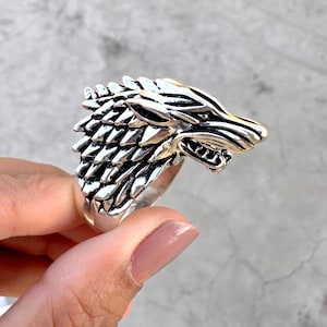May include: A silver ring with a wolf head design. The wolf's head is intricately detailed, showing fur and teeth. The ring is held in a hand, highlighting its size and design. The ring is made of a silver-colored metal.