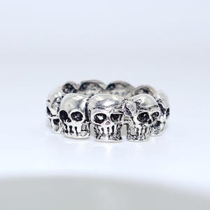 Puede incluir: Un anillo plateado con un patrón repetido de caras de calaveras. Las calaveras tienen rasgos faciales detallados, incluyendo cuencas de los ojos, narices y dientes. El anillo es de metal y tiene un acabado pulido, adecuado para joyería gótica o alternativa.