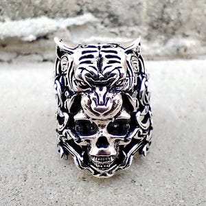 Puede incluir: Anillo plateado con un diseño detallado de la cara de un tigre sobre una calavera. El anillo presenta patrones intrincados y una estética gótica, adecuado para quienes aprecian la joyería audaz.