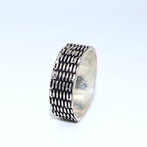 Puede incluir: Un anillo plateado con un patrón texturizado de pequeñas formas alargadas. El anillo tiene un diseño de banda con una serie de impresiones ovaladas repetidas. El anillo está sobre un fondo blanco, resaltando su brillo metálico.
