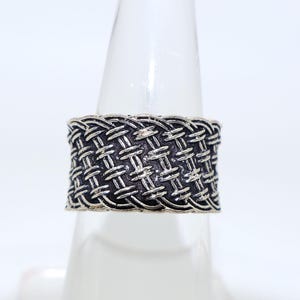 Puede incluir: Un anillo ancho de plata con un diseño de cesta tejida. El anillo está hecho de un metal plateado brillante y presenta un patrón detallado de líneas entrelazadas, creando una superficie texturizada. El anillo se muestra sobre un soporte cónico transparente.