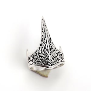 Könnte beinhalten: Ein silberfarbener Fingerring mit einem aufwendigen, gravierten Design. Der Ring hat ein hohes, spitzes zentrales Element und detaillierte Muster auf seiner Oberfläche. Der Ring ist vor einem weißen Hintergrund platziert, der seine komplizierten Details hervorhebt.