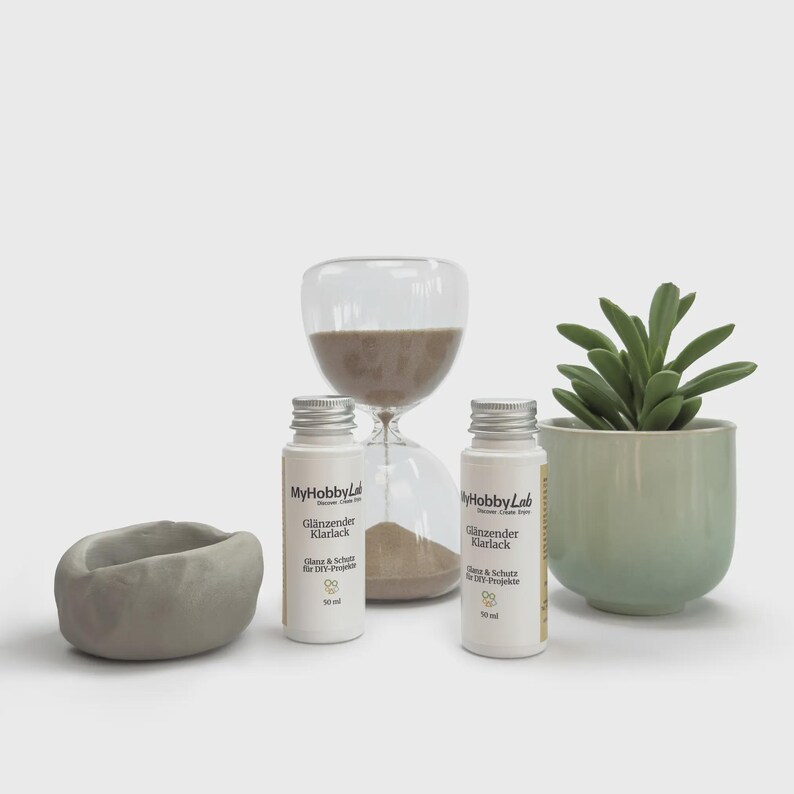 May include: A still life arrangement featuring two bottles of "Gl&auml;nzender Klarlack" with silver caps, a clear hourglass, a small green succulent in a light green pot, and a gray, oval-shaped container.