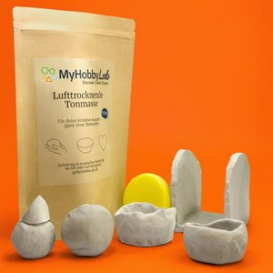 May include: A package of air-drying clay with the text "MyHobbyLab" and "Lufttrocknende Tonmasse". Several small, gray clay sculptures and a yellow disc are arranged in front of the package. The background is orange.