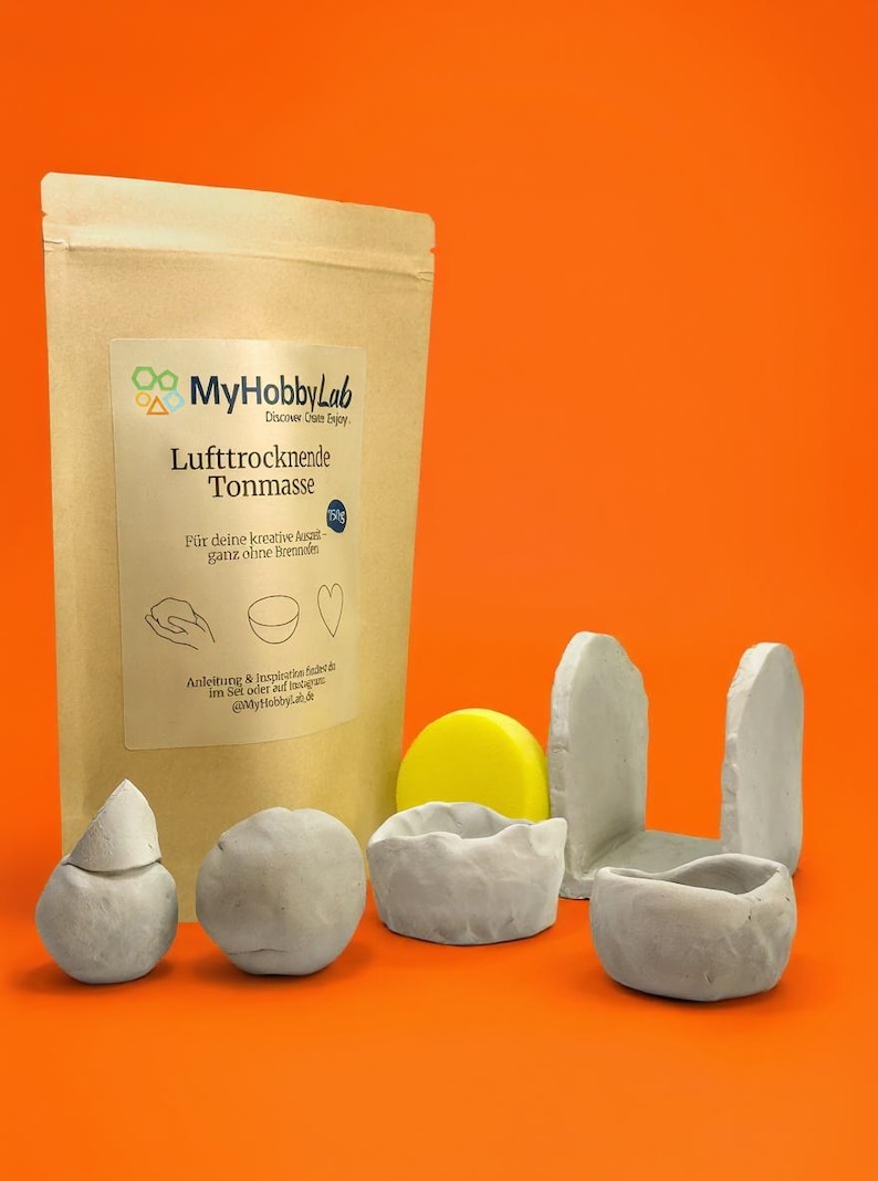 May include: A package of air-drying clay with the text "MyHobbyLab" and "Lufttrocknende Tonmasse". Several small, gray clay sculptures and a yellow disc are arranged in front of the package. The background is orange.