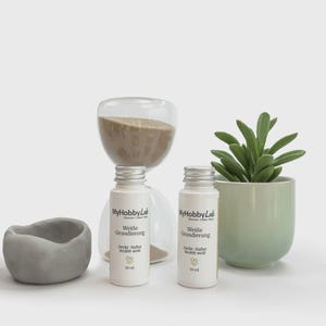 May include: A still-life composition featuring two white bottles with silver caps, labeled "Wei&szlig;e Grundierung" and "MyHobbyLab." An hourglass, a small succulent in a green pot, and a gray, rounded container are also present.