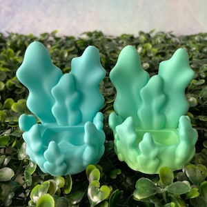 May include: Two decorative teal and mint green candle holders. Each holder features a stylized, organic design resembling aquatic plants. The holders are placed on a bed of green foliage, creating a natural aesthetic.