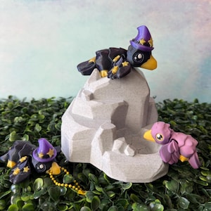 May include: A charming display of bird figurines. Two black birds, sporting yellow beaks and purple wizard hats with stars, perch on a grey rock. A purple bird with a yellow beak is also present. The setting is a backdrop of green foliage.