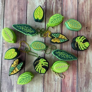 Op de afbeelding: Een verzameling groene en gele blad-vormige sleutelhangers en bedels. De bladeren hebben verschillende ontwerpen en zijn bevestigd aan kleine metalen kogelkettingen. De achtergrond is een houten oppervlak met verticale planken.