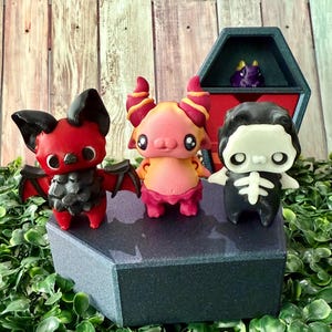 May include: Three colorful, whimsical figurines: a red bat with black wings, a pink and orange creature with horns, and a black skeleton figure. They are displayed on a dark gray coffin-shaped box, with a small purple dragon in a coffin-shaped box in the background.