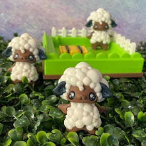 May include: Three 3D-printed sheep figurines with white fluffy wool, brown faces and limbs, and dark blue ears. One sheep is in a green pen with a white fence and yellow objects. The figurines are set on a bed of green foliage.