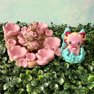 Puede incluir: Dos figuras caprichosas: una marrón con detalles de flores rosas, la otra rosa con detalles amarillos y rojos. La primera está rodeada de flores rosas, la segunda está en una estructura azul. Ambas tienen ojos grandes.