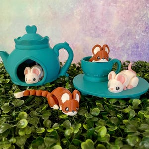 Puede incluir: Un juego de tetera y taza de té turquesa caprichoso con figuras de ratón en miniatura. La tetera tiene una tapa en forma de corazón y un ratón en el interior. La taza de té contiene un ratón, y otro ratón está en el platillo.