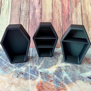 Könnte beinhalten: Drei schwarze, sargförmige Regale. Die Regale bestehen aus einem mattschwarzen Material und haben eine sechseckige Form. Das Regal in der Mitte hat zwei Regalböden. Die beiden anderen Regale haben ein offenes Design. Die Regale werden auf einer Marmoroberfläche ausgestellt.