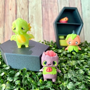 May include: A selection of quirky figurines in vibrant colours. A green, two-headed creature with wings is on a dark grey platform. A purple figure with a brain-like head and a green shirt is in front. A pumpkin-headed figure and a small orange creature are in the background.