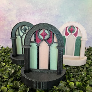 May include: Three decorative arched window ornaments. The windows feature a stained-glass design with a central starburst and vertical panels in shades of pink, green, and white. The frames are black and white, and the bases are black.