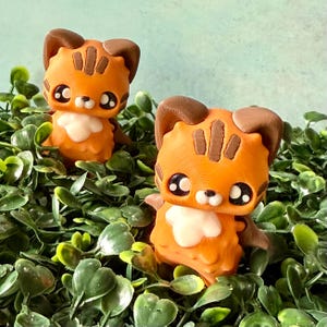 May include: Two orange and brown cat figurines with large black eyes and white chest details are nestled among green foliage. The cat figurines have a cartoonish design with textured details. The background is a light blue color.