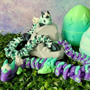 May include: A collection of colorful, articulated dragon toys in shades of purple, green, and black. One dragon rests on a gray rock formation, while others are coiled or extended. Two green and white egg-shaped containers are also present.