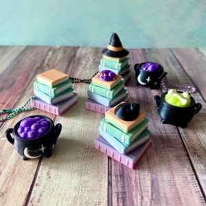 Può includere: Ciondoli in miniatura a tema Halloween con pile di libri, cappelli da strega, calderoni e gatti neri. I libri sono in colori pastello e i calderoni contengono sostanze viola e verdi. Su una superficie di legno.