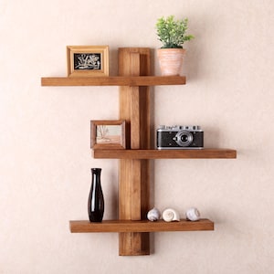 May include: A wooden wall shelf unit with three shelves, displaying a framed picture, a potted plant, a vintage camera, a black vase, and seashells. The wood has a natural finish. The unit is mounted on a wall.