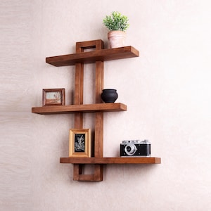 May include: A dark brown wooden wall shelf with three horizontal shelves and vertical supports. The shelves hold a small potted plant, a black ceramic vase, two framed pictures, and a vintage camera.