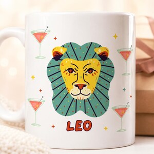 Puede incluir: Taza de cerámica blanca con un diseño de cabeza de león estilizada, con una melena turquesa y cara amarilla. La palabra "LEO" está impresa en naranja. La taza está decorada con copas de cóctel y estrellas, para los entusiastas de la astrología.