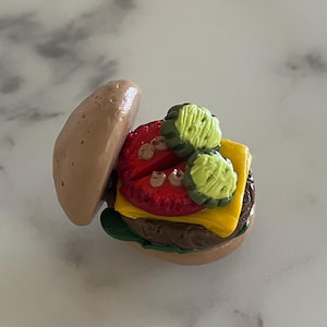 May include: A miniature burger with a tan bun, a red tomato, yellow cheese, green pickles, and a brown patty. The burger is partially open, revealing the ingredients. The burger is a food-themed item.