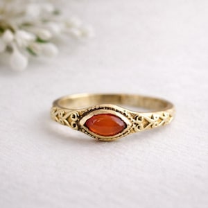 May include: A gold-colored ring with an oval, orange gemstone at the center. The band features an ornate, swirling design. The ring is set against a soft, white background, highlighting the jewelry's details.