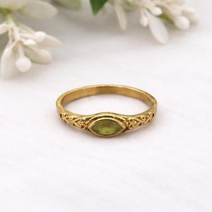 May include: A gold ring with a light green, oval gemstone. The ring has an ornate, openwork design on the band. The ring is set against a white background, with soft floral accents in the upper corners.
