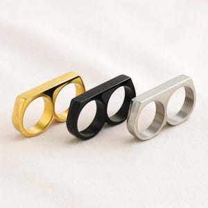 Peut inclure: Trois bagues à deux doigts, dorées, noires et argentées. Chaque bague comporte deux ouvertures circulaires reliées par une barre rectangulaire. Les bagues sont présentées sur une surface blanche.