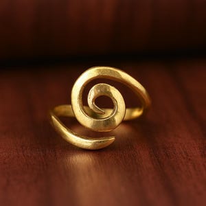 May include: A gold-colored ring with a spiral design. The ring features a swirling, open-ended spiral shape, with a smooth, polished surface. The ring is set against a dark brown background, highlighting its metallic sheen.