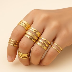 May include: A hand wearing multiple gold rings. The rings vary in design, including smooth bands, textured surfaces, and stacked ring styles. The rings are a bright, shiny gold color.