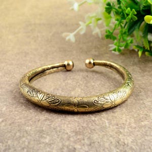 May include: A gold-colored cuff bracelet with an open design. The bracelet features an embossed floral pattern along its surface and rounded end caps. The jewelry is displayed on a neutral-toned surface.