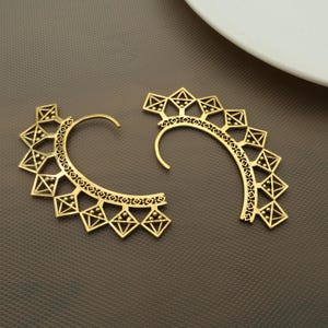 May include: Two gold-colored ear cuffs with geometric designs. Each cuff features a curved shape with a series of diamond and triangle cutouts along the edges. The earrings are displayed on a brown surface.