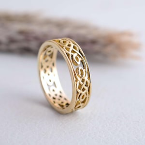 May include: A gold-colored ring with an intricate Celtic knot design. The ring features an openwork pattern with interwoven lines and curves. The band is approximately 0.2 inches wide, with a polished finish. The ring is set against a soft, neutral background.