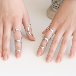 Puede incluir: Anillos plateados en varios diseños. Uno es una banda simple, mientras que otros presentan diseños alargados y calados que cubren múltiples segmentos de los dedos. Los anillos se muestran sobre un fondo claro.