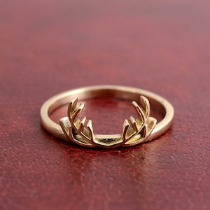 May include: A gold-colored ring featuring a detailed antler design. The ring is set against a red background, highlighting the intricate details of the antlers. The ring is a piece of jewelry.