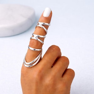 May include: A silver finger ring set. The ring set includes a series of connected rings that cover the finger. The rings have a modern, geometric design. The ring set is worn on a hand with a white painted fingernail.
