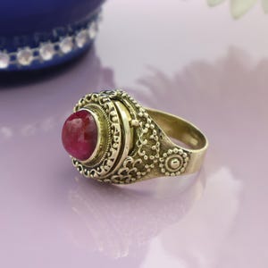 May include: A vintage-style ring with a large, oval, red gemstone set in an ornate, gold-toned metal band. The ring features detailed filigree and scrollwork around the gemstone and on the band, showcasing a classic, antique aesthetic.