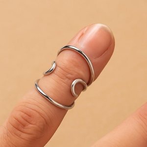 May include: A silver-colored finger ring with a unique design. The ring wraps around the finger with curved and circular elements. The ring is shown on a person's finger against a neutral background.