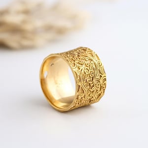 May include: A gold-colored ring with an intricate floral pattern. The ring has a wide band and a polished finish. The floral design covers the entire surface of the ring, creating a textured appearance. The ring is set against a white background.