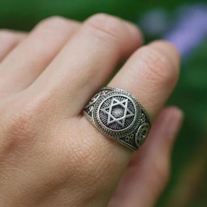 May include: Silver ring with a prominent Star of David design at its centre. The ring has detailed patterns around the star and along the band, suggesting a vintage or antique aesthetic. The ring is worn on a finger.